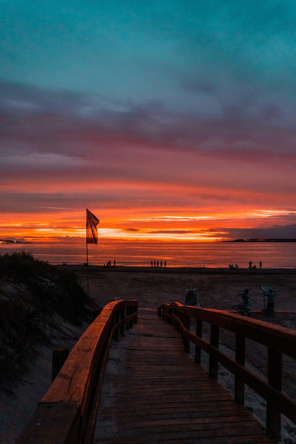 Bajada de madera a la playa de Punta del Este en un atardecer de verano, con la bandera y gente en la orilla
