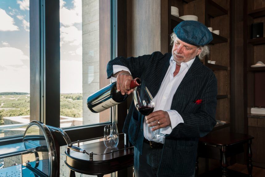 Francis Mallmann sirviendo vino frente al ventanal, con las viñas de fondo