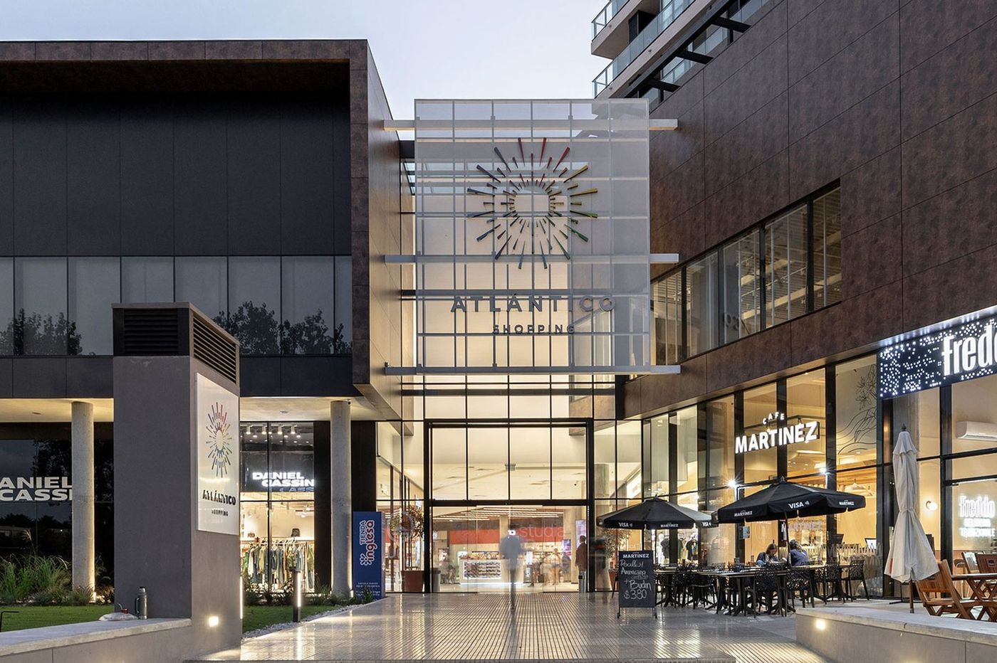 Acceso peatonal del shopping al anochecer, con los cafés y locales encendidos