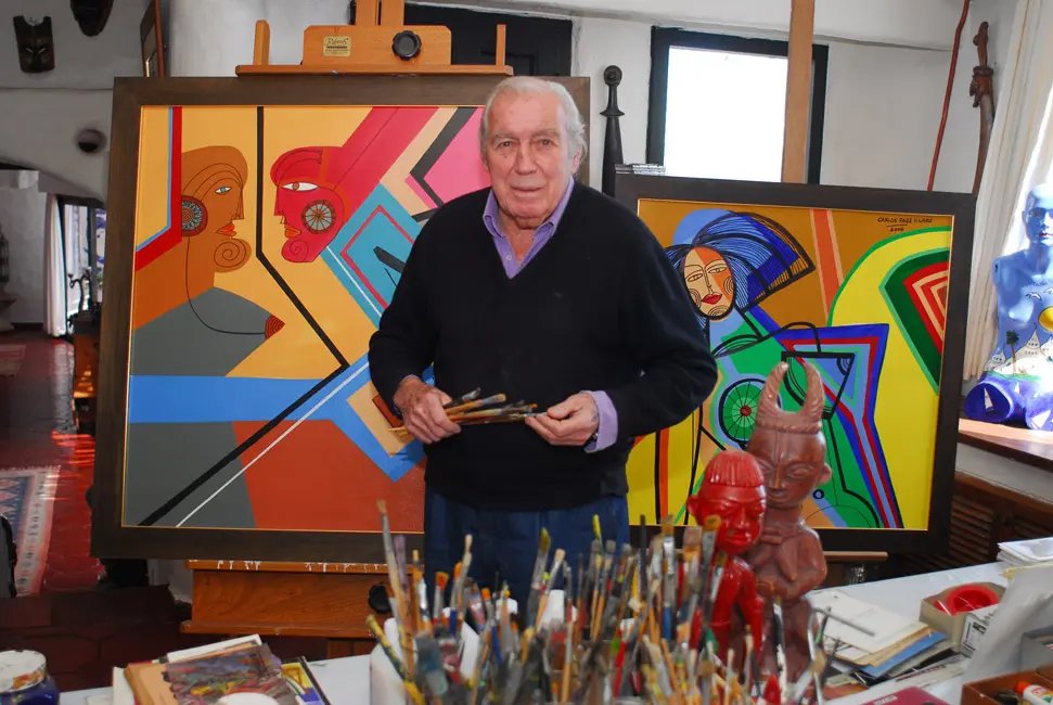 Carlos Páez Vilaró em seu ateliê em Casapueblo, com pincéis na mão e dois de seus quadros ao fundo