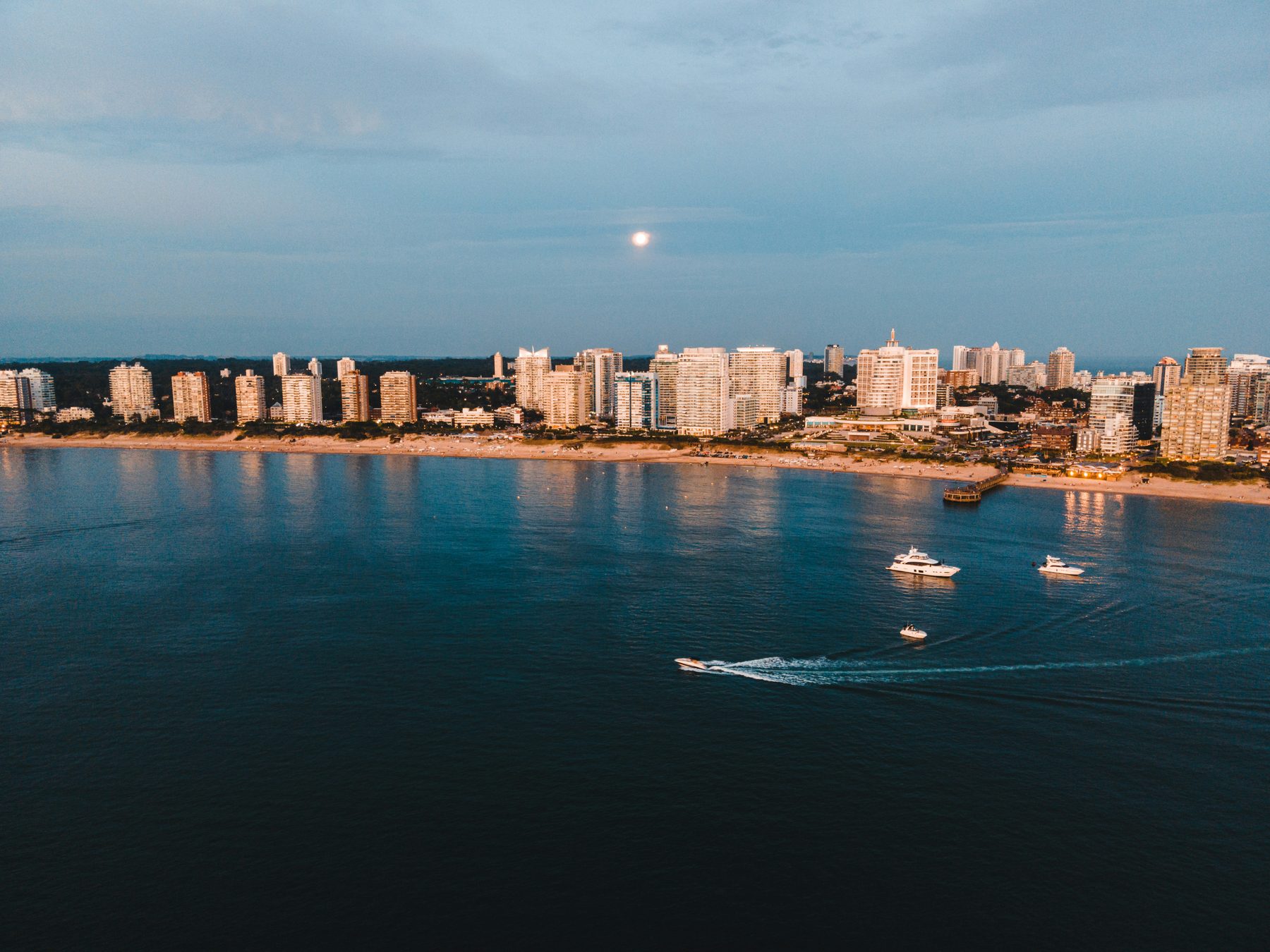 Vista aérea de la Playa Mansa de Punta del Este al anochecer, con yates fondeados y la luna sobre las torres