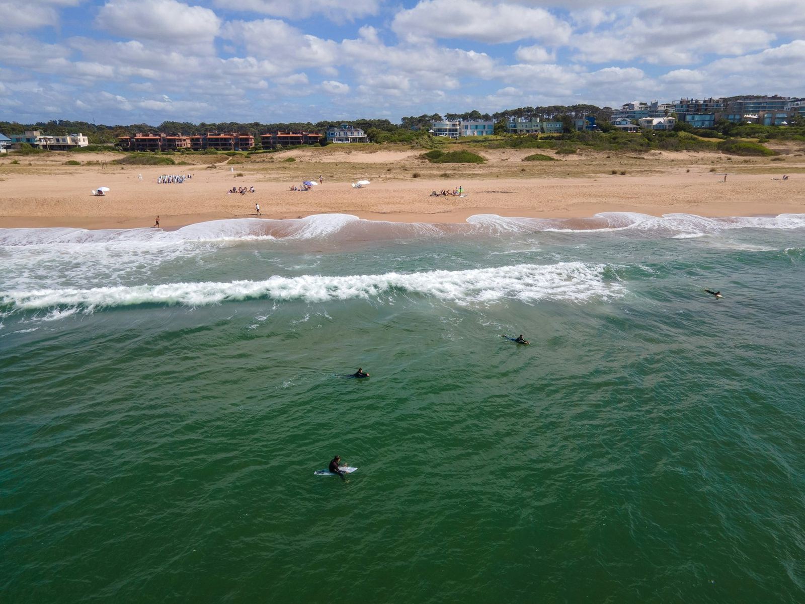 Vista aérea de surfistas en el mar frente a la playa, con las casas sobre los médanos